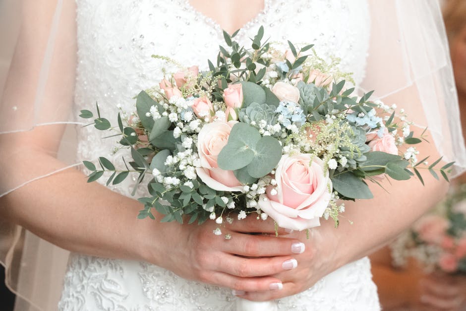 A bride holding a delicate bouquet arrangement featuring soft pink roses, pale blue hydrangeas, sprigs of baby's breath, and eucalyptus leaves. The flowers are fresh, with velvety petals and sturdy gr