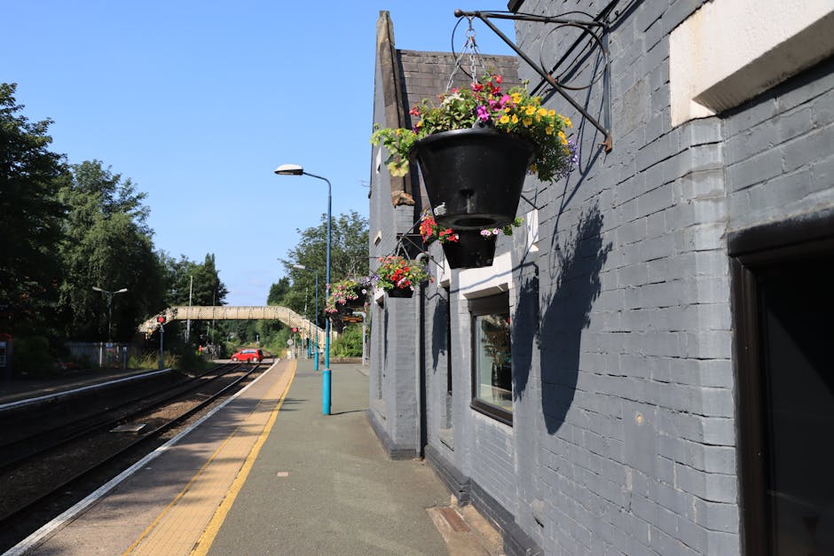A flower delivery scene featuring a large black hanging flower basket filled with vibrant pink, yellow, purple, and white blossoms, attached to the grey brick exterior wall of a building. The basket c
