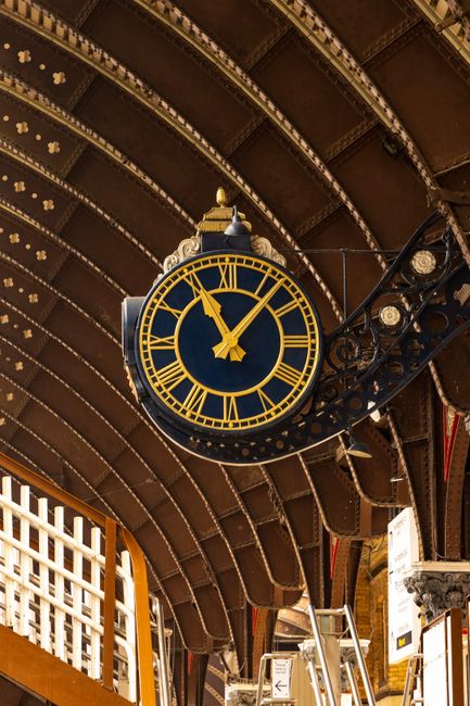 A large, vintage-style clock with a black frame and gold numerals and hands is mounted on an ornate wrought iron bracket. The clock displays a time of approximately 10:09. The background features a cu