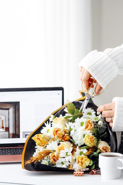 A close-up of a person's hand in a white knitted sleeve, holding a pair of scissors and carefully trimming a vibrant floral bouquet placed on a desk. The bouquet includes soft yellow roses, white dais