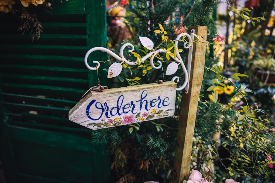 A decorative wooden garden sign with the words 'Order here' painted in blue cursive, hanging from an ornate white metal bracket attached to a yellow wooden post. The sign is surrounded by lush green f