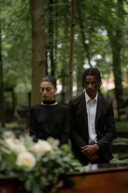 Two individuals with solemn expressions stand outdoors surrounded by lush green trees during daylight, dressed in dark, formal attire; the woman wears a black top with a choker and the man dons a blac