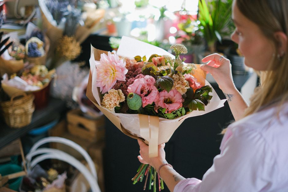 A woman holding a bouquet of fresh flowers in a florist shop. The bouquet includes pink dahlias, pale pink roses, dark green foliage, and other pastel-colored blooms, all arranged in light-colored wra